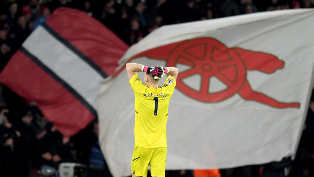 Aaron Ramsdale stands with hands on head. Arsenal flags wave in front of him.